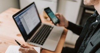 Person arbeitet an einem Laptop, hält in der einen Hand ein Smartphone und macht gleichzeitig handschriftliche Notizen.